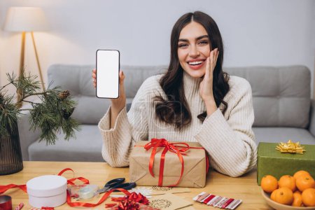 Photo for Beautiful happy woman doing New Year or Christmas gift wrapping while holding smart phone background with blank screen with copy space or mock up at home - Royalty Free Image
