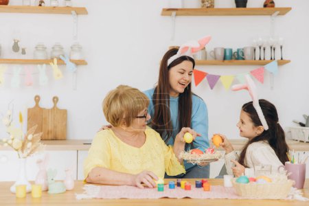 Foto de ¡Feliz Pascua! Tres generaciones de mujeres, madre feliz con hija y abuela pintando huevos de colores y pasándola bien juntas en la cocina - Imagen libre de derechos