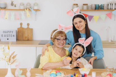 Photo for Happy Easter! Three generations of women, happy mother with daughter and grandmother painting colorful eggs and having a good time together in the kitchen - Royalty Free Image