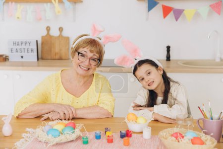 Téléchargez les photos : Bonne grand-mère et petite-fille dans les oreilles de lapin de Pâques ayant oeuf tapant avec des oeufs de Pâques à la maison moderne dans le jour ensoleillé du printemps - en image libre de droit