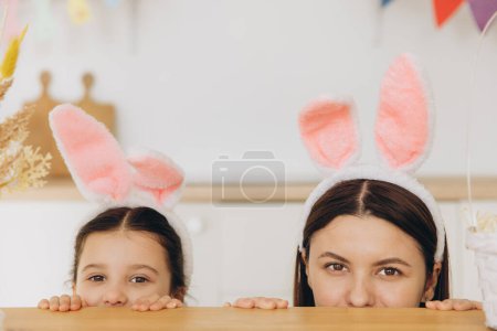 Foto de ¡Feliz Pascua! Joven madre y linda hija con orejas de conejo mientras se esconde detrás de la mesa y divertirse en la cocina en casa - Imagen libre de derechos