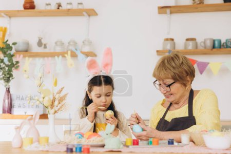 Foto de ¡Feliz Pascua! Una abuela y su nieta pintando huevos de Pascua. Familia feliz preparándose para la Pascua. Linda niña pequeña con orejas de conejo en el día de Pascua
. - Imagen libre de derechos