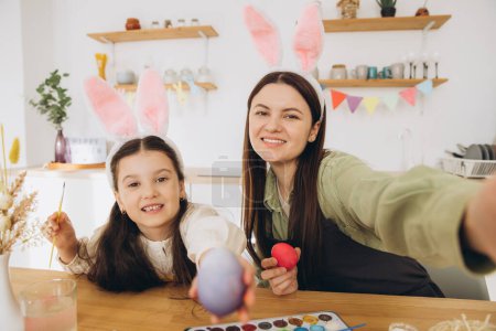 Téléchargez les photos : Mère et sa fille peignent des œufs et font un selfie ou un appel vidéo. Bonne famille se préparant pour Pâques. Petite fille mignonne portant des oreilles de lapin. - en image libre de droit