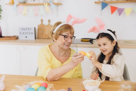 Foto de Feliz abuela y nieta en orejas de conejo de Pascua teniendo huevo golpeando con huevos de Pascua en casa moderna en soleado día de primavera - Imagen libre de derechos
