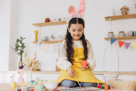 Foto de ¡Feliz Pascua! Una hermosa niña sentada en la mesa con decorationes y pintando huevos de colores. Linda niña con orejas de conejo en el día de Pascua. - Imagen libre de derechos