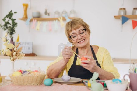 Téléchargez les photos : Joyeux Pâques ! Mignon retraité vieille femme peint oeufs de Pâques sur la cuisine à la maison. Oeufs de Pâques bricolage - en image libre de droit