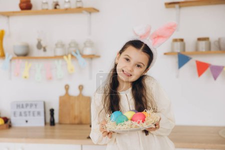 Foto de Retrato de linda niña feliz con orejas de conejito de felpa y delantal sosteniendo cesta de mimbre llena de huevos de colores mult decorados creativamente de Pascua, mientras que de pie cocina con decoraciones festivas - Imagen libre de derechos