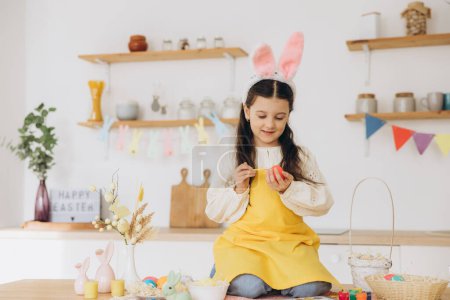 Téléchargez les photos : Joyeux Pâques ! Une belle fillette assise sur la table avec des décorations et peignant des œufs colorés. Mignon enfant fille portant des oreilles de lapin le jour de Pâques. - en image libre de droit
