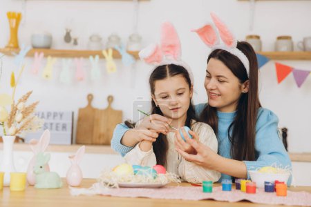 Foto de Madre y su hija pintando huevos. Familia feliz preparándose para la Pascua. Linda niña pequeña con orejas de conejito. - Imagen libre de derechos