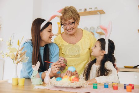 Foto de ¡Feliz Pascua! Tres generaciones de mujeres, madre feliz con hija y abuela pintando huevos de Pascua y pasando un buen rato juntas en la cocina - Imagen libre de derechos