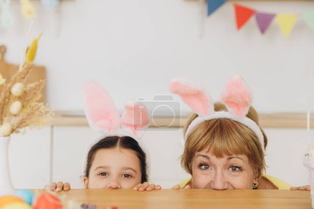 Téléchargez les photos : Joyeux Pâques ! Grand-mère et mignonne petite-fille portant des oreilles de lapin tout en se cachant derrière la table et avoir du plaisir dans la cuisine à la maison - en image libre de droit