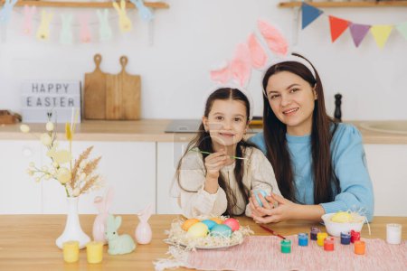 Foto de Madre y su hija pintando huevos. Familia feliz preparándose para la Pascua. Linda niña pequeña con orejas de conejito. - Imagen libre de derechos