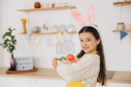 Photo for Portrait of cute happy little girl wearing plush bunny ears and apron holding wicker basket full of Easter creatively decorated mult colored eggs while standing kitchen with festive decorations - Royalty Free Image