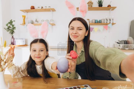Photo for Mother and her daughter painting eggs and making selfie or video call. Happy family preparing for Easter. Cute little child girl wearing bunny ears. - Royalty Free Image