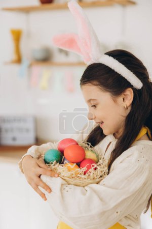 Photo for Portrait of cute happy little girl wearing plush bunny ears and apron holding wicker basket full of Easter creatively decorated mult colored eggs while standing kitchen with festive decorations - Royalty Free Image
