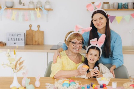 Foto de ¡Feliz Pascua! Tres generaciones de mujeres, madre feliz con hija y abuela pintando huevos de colores y pasándola bien juntas en la cocina - Imagen libre de derechos