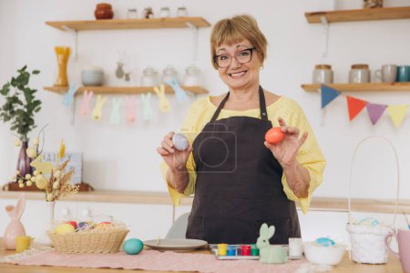 Foto de ¡Feliz Pascua! Linda anciana jubilada pintó huevos de Pascua en la cocina en casa. Artesanía DIY huevos de Pascua - Imagen libre de derechos