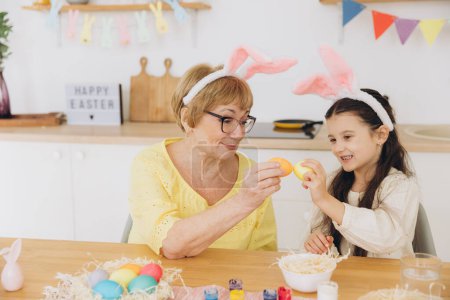Téléchargez les photos : Bonne grand-mère et petite-fille dans les oreilles de lapin de Pâques ayant oeuf tapant avec des oeufs de Pâques à la maison moderne dans le jour ensoleillé du printemps - en image libre de droit