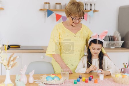 Foto de ¡Feliz Pascua! Una abuela y su nieta pintando huevos de Pascua. Familia feliz preparándose para la Pascua. Linda niña pequeña con orejas de conejo en el día de Pascua
. - Imagen libre de derechos