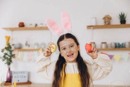 Foto de ¡Feliz Pascua! Linda niña sosteniendo coloridos huevos de Pascua y divertirse en la cocina en casa. Niña con orejas de conejo el día de Pascua. - Imagen libre de derechos