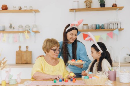 Téléchargez les photos : Joyeux Pâques ! Trois générations de femmes, mère heureuse avec fille et grand-mère peignant des œufs colorés et s'amusant ensemble dans la cuisine - en image libre de droit