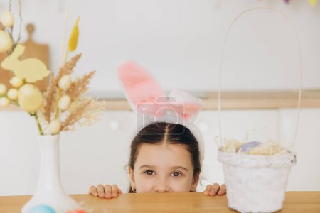 Téléchargez les photos : Joyeux Pâques ! Petite fille mignonne portant des oreilles de lapin tout en se cachant derrière la table et amusez-vous dans la cuisine à la maison - en image libre de droit