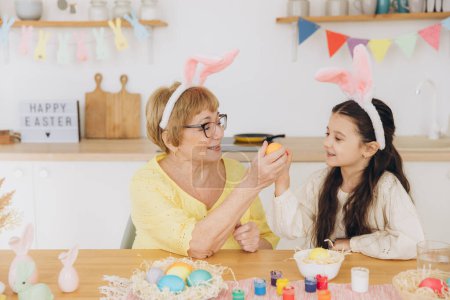 Foto de Feliz abuela y nieta en orejas de conejo de Pascua teniendo huevo golpeando con huevos de Pascua en casa moderna en soleado día de primavera - Imagen libre de derechos
