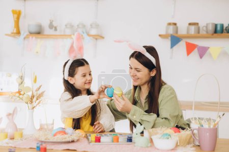 Téléchargez les photos : Mère et sa fille s'embrassent et tiennent des œufs colorés. Bonne famille se préparant pour Pâques. Petite fille mignonne portant des oreilles de lapin. - en image libre de droit