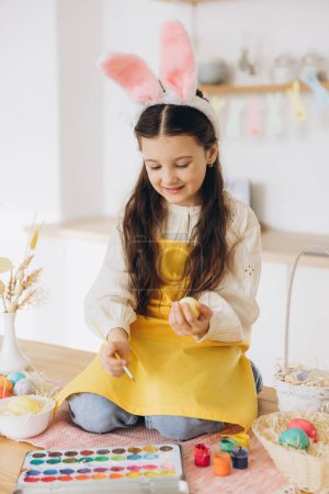 Foto de ¡Feliz Pascua! Una hermosa niña sentada en la mesa con decorationes y pintando huevos de colores. Linda niña con orejas de conejo en el día de Pascua. - Imagen libre de derechos