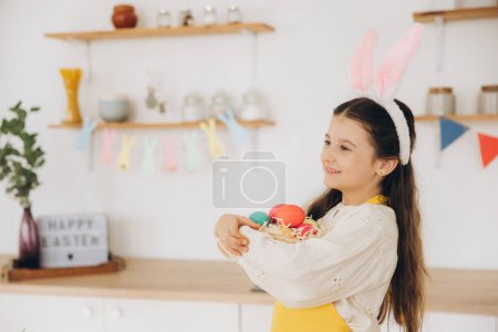 Foto de Retrato de linda niña feliz con orejas de conejito de felpa y delantal sosteniendo cesta de mimbre llena de huevos de colores mult decorados creativamente de Pascua, mientras que de pie cocina con decoraciones festivas - Imagen libre de derechos