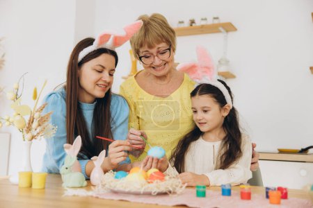 Foto de ¡Feliz Pascua! Tres generaciones de mujeres, madre feliz con hija y abuela pintando huevos de Pascua y pasando un buen rato juntas en la cocina - Imagen libre de derechos