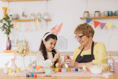 Photo for Happy easter! A grandmother and her granddaughter painting Easter eggs. Happy family preparing for Easter. Cute little child girl wearing bunny ears on Easter day. - Royalty Free Image