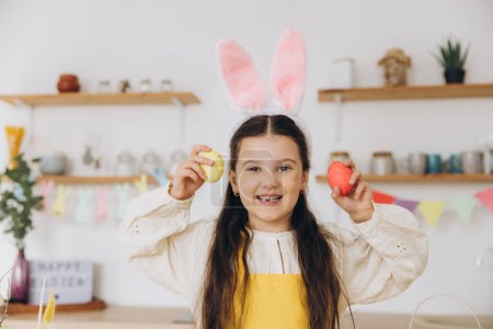 Téléchargez les photos : Joyeux Pâques ! Petite fille mignonne tenant des œufs de Pâques colorés et s'amusant sur la cuisine à la maison. Enfant fille portant des oreilles de lapin le jour de Pâques. - en image libre de droit