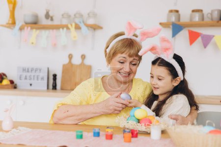 Photo for Happy easter! A grandmother and her granddaughter painting Easter eggs. Happy family preparing for Easter. Cute little child girl wearing bunny ears on Easter day. - Royalty Free Image