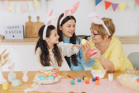 Photo for Happy Easter! Three generations of women, happy mother with daughter and grandmother painting colorful eggs and having a good time together in the kitchen - Royalty Free Image