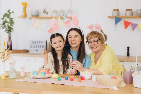 Téléchargez les photos : Joyeux Pâques ! Trois générations de femmes, mère heureuse avec fille et grand-mère peignant des œufs colorés et s'amusant ensemble dans la cuisine - en image libre de droit