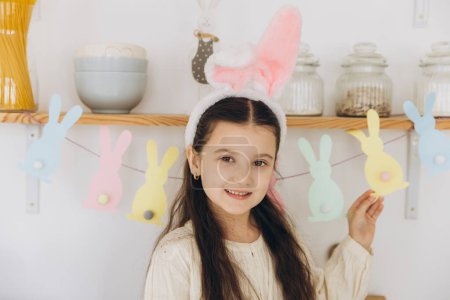 Téléchargez les photos : Joyeux Pâques ! Portrait de mignonne petite fille heureuse portant des oreilles de lapin en peluche tout en restant debout cuisine avec des décorations festives - en image libre de droit