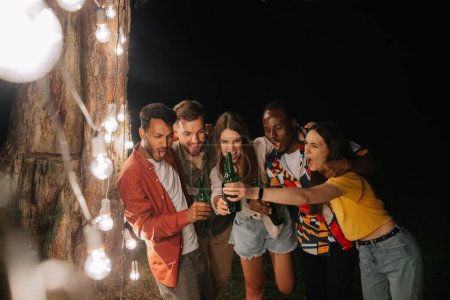 Téléchargez les photos : Une compagnie d'amis multiraciaux buvant de la bière à la fête, faisant des visages près des lampes suspendues - en image libre de droit