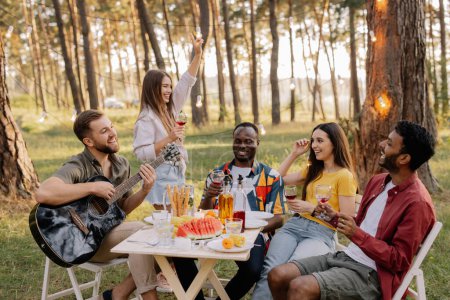 Téléchargez les photos : Rencontre d'amis multiraciaux jouant de la guitare, chantant, dînant et buvant du vin pendant la fête dans la forêt - en image libre de droit