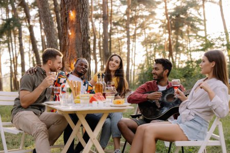 Foto de Encuentro de grupo multirracial de amigos tocando la guitarra, cantando, cenando y bebiendo vino durante la fiesta en el bosque - Imagen libre de derechos