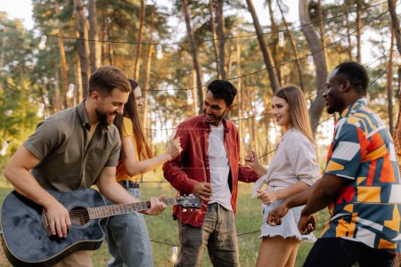 Téléchargez les photos : Groupe multiracial de personnes, homme barbu hipster jouer de la guitare et des amis danser, chanter et s'amuser - en image libre de droit