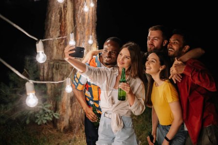 Foto de Un grupo de amigos multirraciales divirtiéndose y bebiendo cerveza se toman selfies cerca de lámparas colgantes por la noche - Imagen libre de derechos