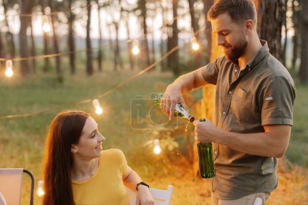 Téléchargez les photos : Un hipster barbu ouvre une bière lors d'une fête entre amis dans une forêt décorée de lampes suspendues - en image libre de droit