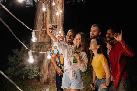 Téléchargez les photos : Un groupe d'amis multiraciaux s'amuser et boire de la bière prennent selfies près des lampes suspendues la nuit - en image libre de droit