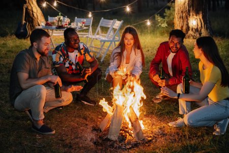 Foto de Amigos multirraciales sentados alrededor del fuego bebiendo cerveza y divirtiéndose en la fiesta - Imagen libre de derechos