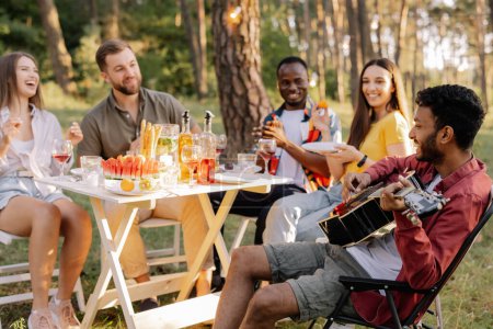 Foto de Grupo multirracial de personas, hipster indio tocando la guitarra y amigos cenando y bebiendo vino durante la fiesta en el bosque - Imagen libre de derechos