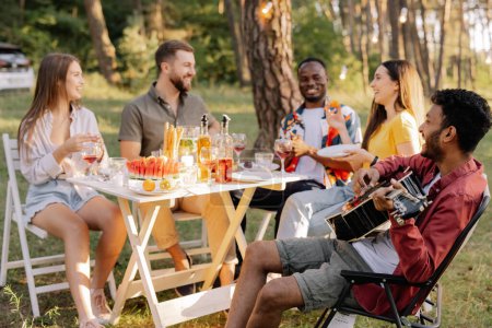 Foto de Grupo multirracial de personas, hipster indio tocando la guitarra y amigos cenando y bebiendo vino durante la fiesta en el bosque - Imagen libre de derechos