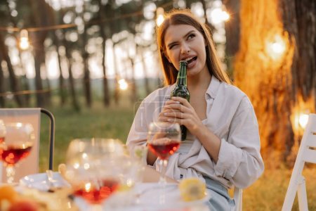 Foto de Mujer hipster divertido abre cerveza con dientes en la fiesta con amigos en el bosque decorado con lámparas colgantes - Imagen libre de derechos