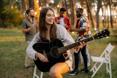 Foto de Hermosa chica hipster tocando la guitarra en el fondo de una cena al aire libre de amigos multirraciales - Imagen libre de derechos