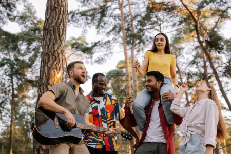Téléchargez les photos : Groupe multiracial de personnes, homme barbu hipster jouer de la guitare et des amis danser, chanter et s'amuser - en image libre de droit